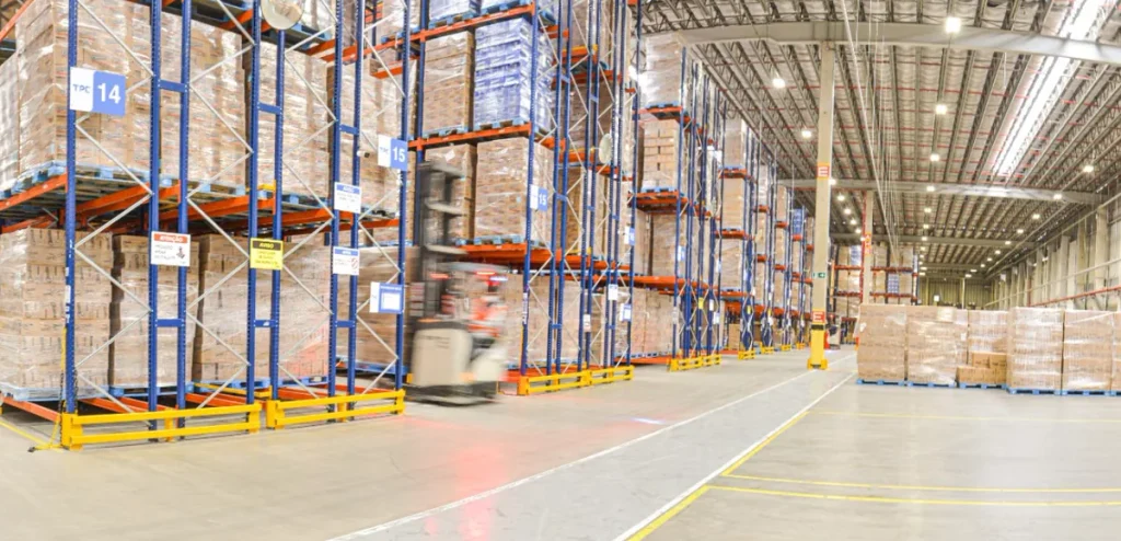 Corredor de centro de distribuição com porta-paletes cheios de caixas e empilhadeira em movimento, mostrando a organização do layout do estoque